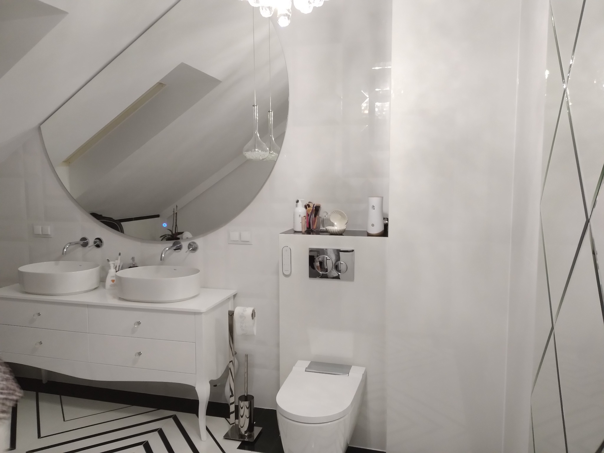 Nowoczesna łazienka w bieli z podwójną umywalką na szafce w stylu retro, okrągłym lustrem i toaletą z chromowanymi akcentami oraz wzorzystą podłogą.