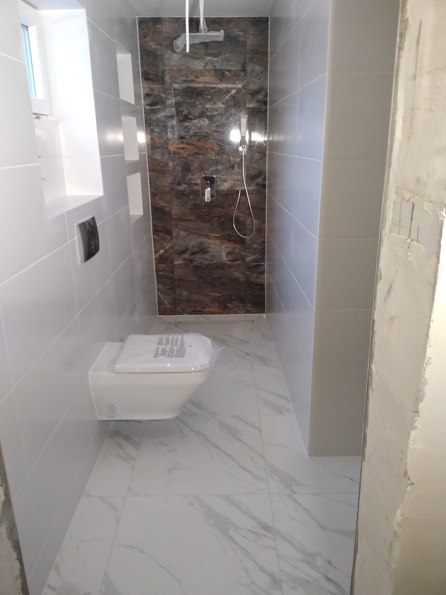 Wąska łazienka po remoncie: białe płytki imitujące marmur na podłodze i ścianach, wisząca toaleta z folią ochronną, prysznic z panelami imitującymi kamień, widoczne niedokończone wykończenie ściany...