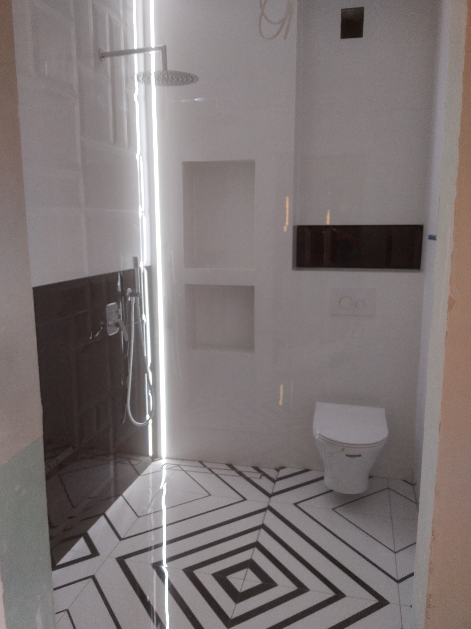 Nowoczesna łazienka z biało-czarną geometryczną podłogą, prysznicem typu deszczownica i wiszącą toaletą; widoczne niedokończone prace wykończeniowe przy ścianach.