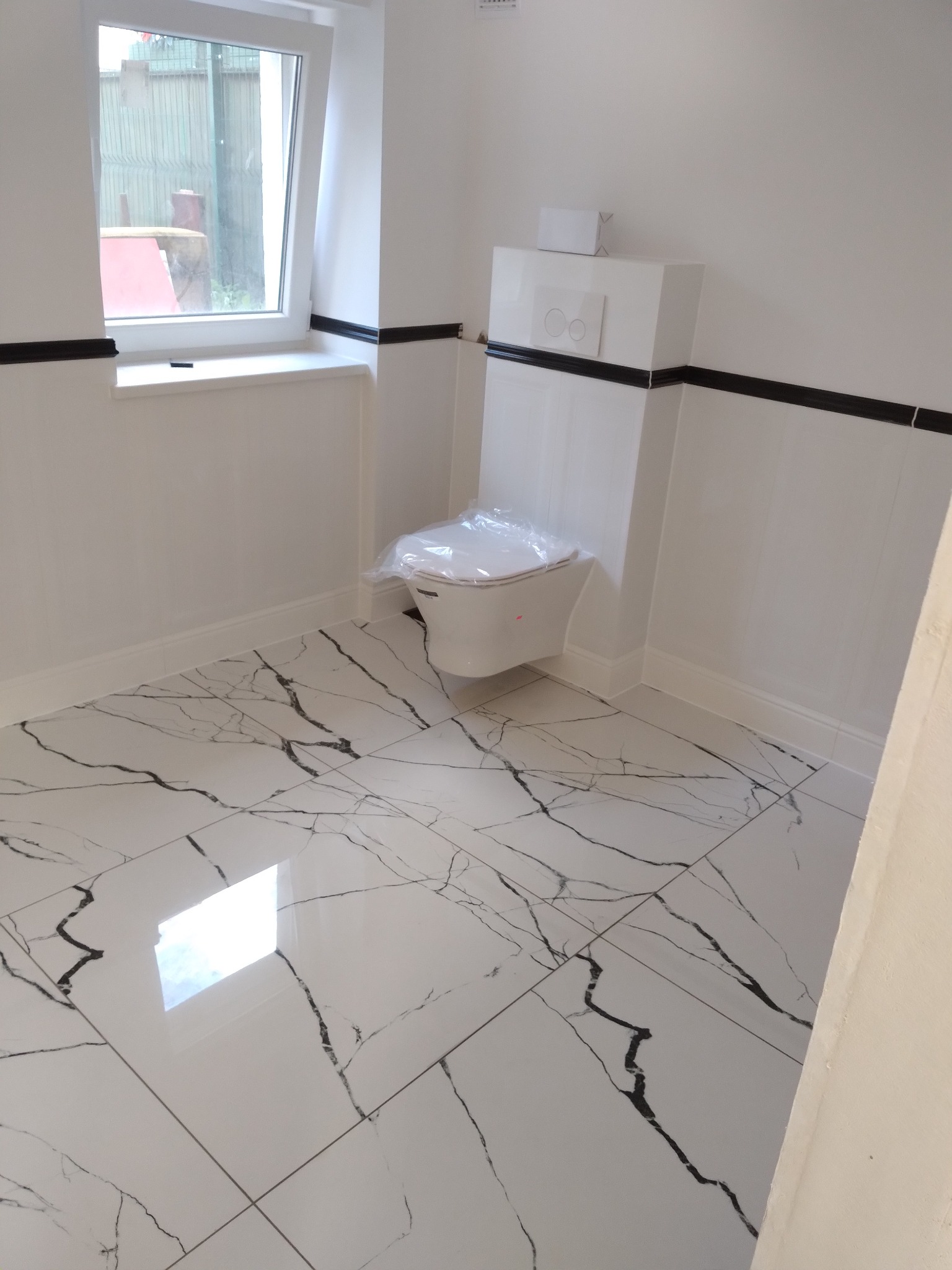 Nowoczesna łazienka z białą armaturą i podłogą z płytek imitujących marmur; toaleta w folii ochronnej, czarne akcenty dekoracyjne.