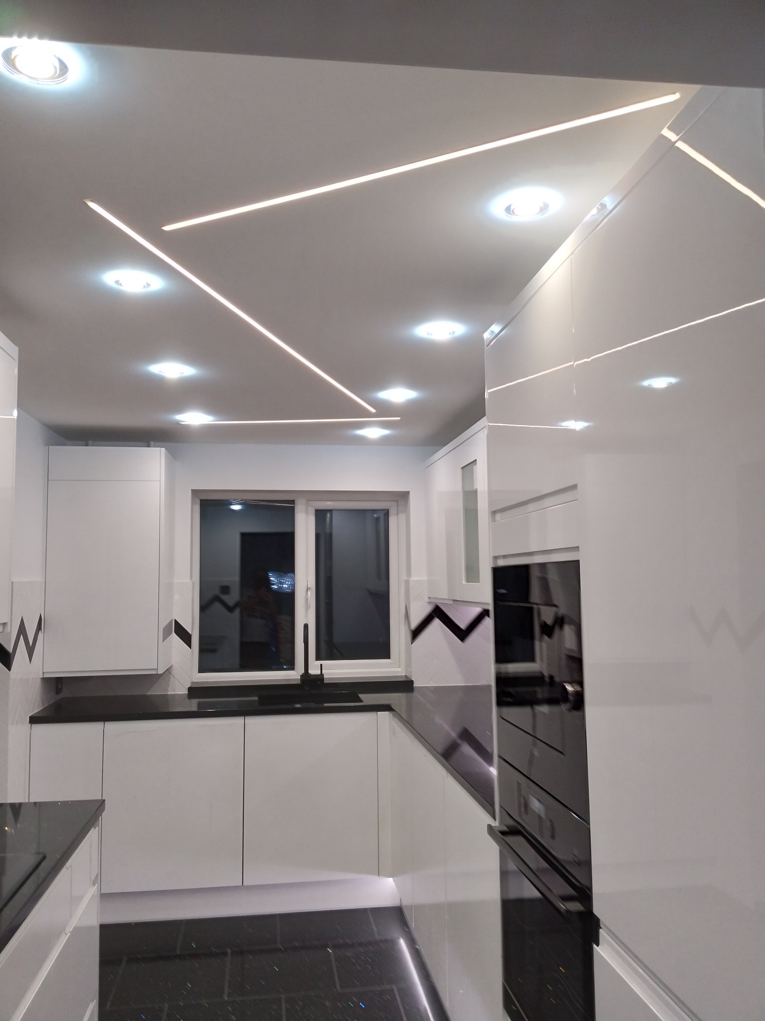 Nowoczesna kuchnia w bieli z czarnym blatem i podłogą, oświetlenie sufitowe LED w geometryczne wzory, okno z czarną ramą i minimalistyczną armaturą.