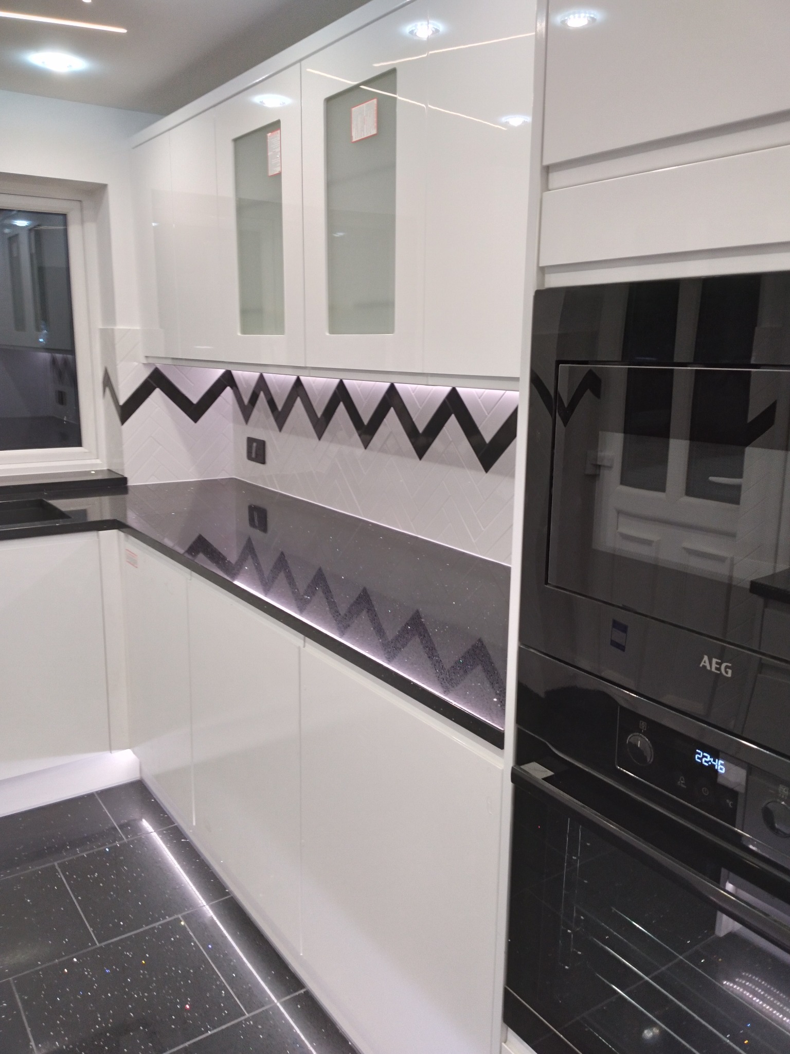 Nowoczesna, czarno-biała kuchnia z błyszczącym, czarnym blatem i podświetleniem LED, wzór zygzak na ścianie, zabudowany piekarnik AEG.