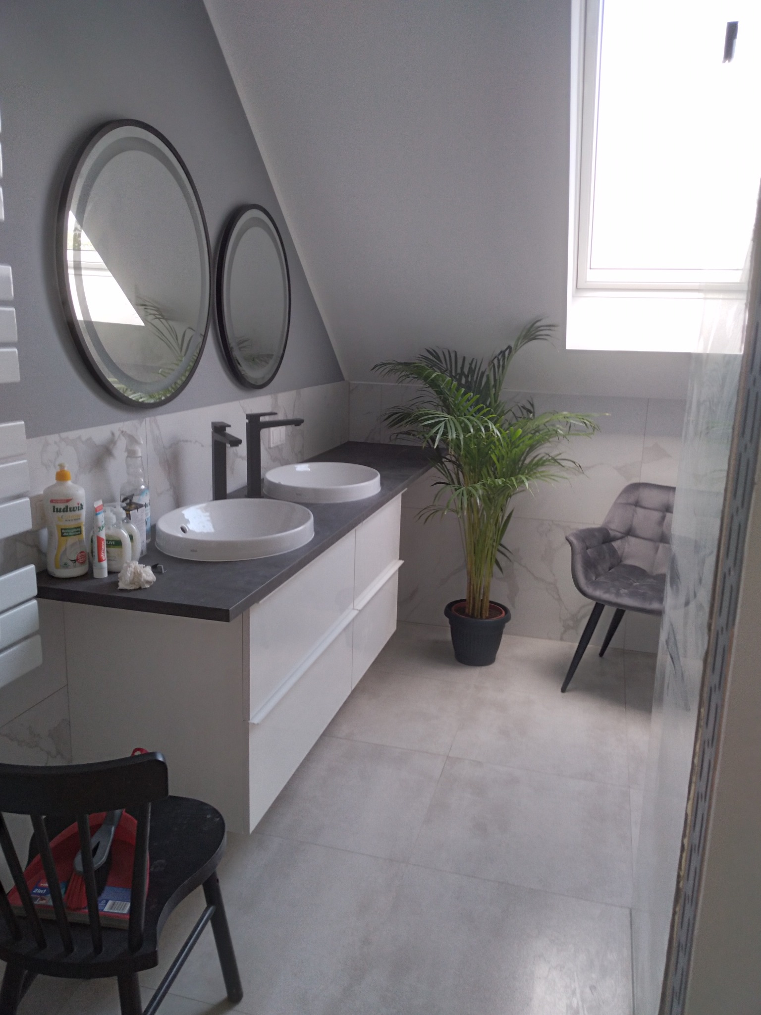 Nowoczesna łazienka na poddaszu z dwoma okrągłymi lustrami, podwójną umywalką na białej szafce z szarym blatem, palmą w doniczce i szarym fotelem; na pierwszym planie czarne krzesło z narzędziami.