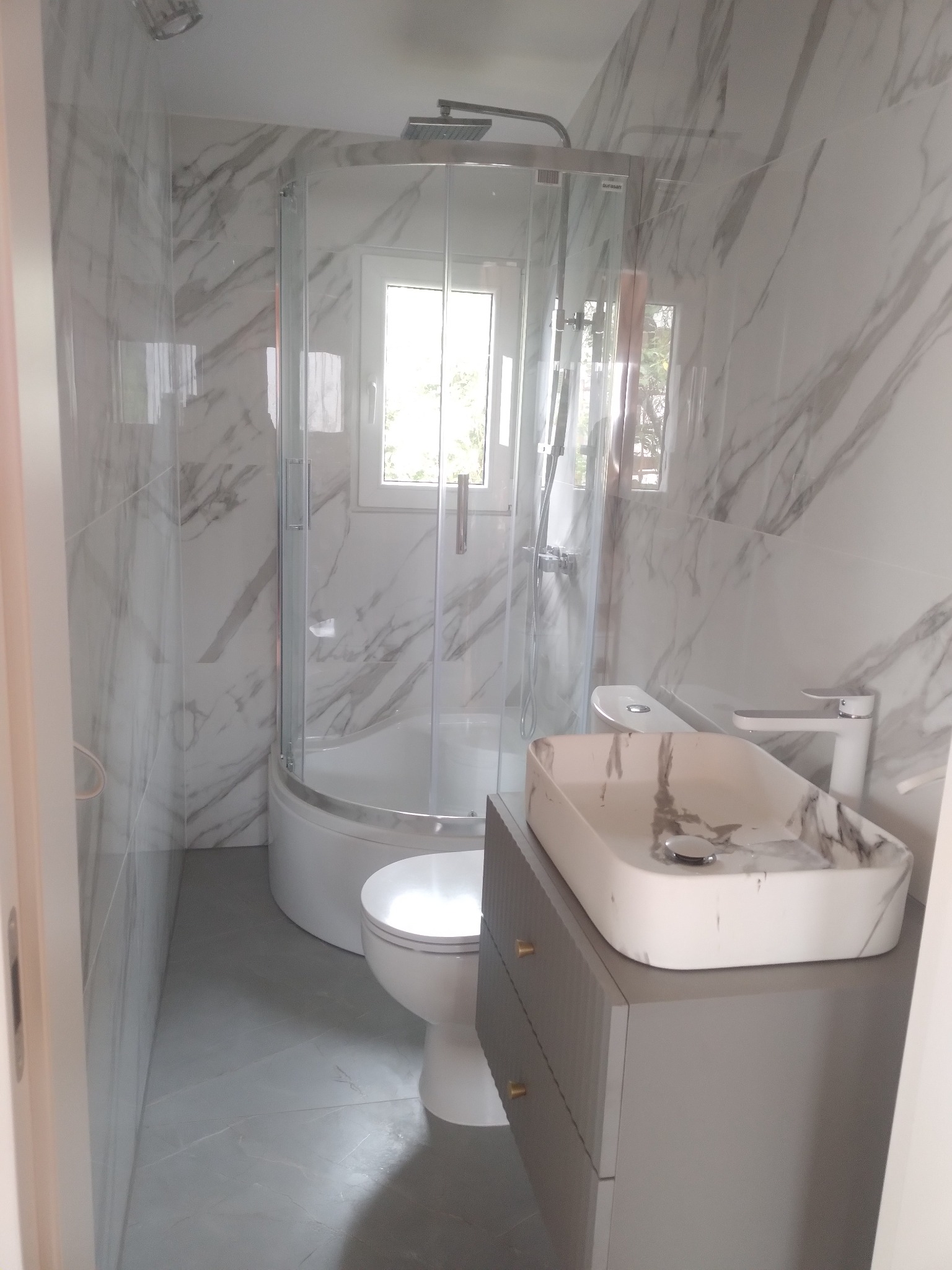 Wykończona łazienka z kabiną prysznicową narożną, toaletą i umywalką nablatową w marmurkowym wzorze, szare płytki podłogowe i marmurowe płytki ścienne.