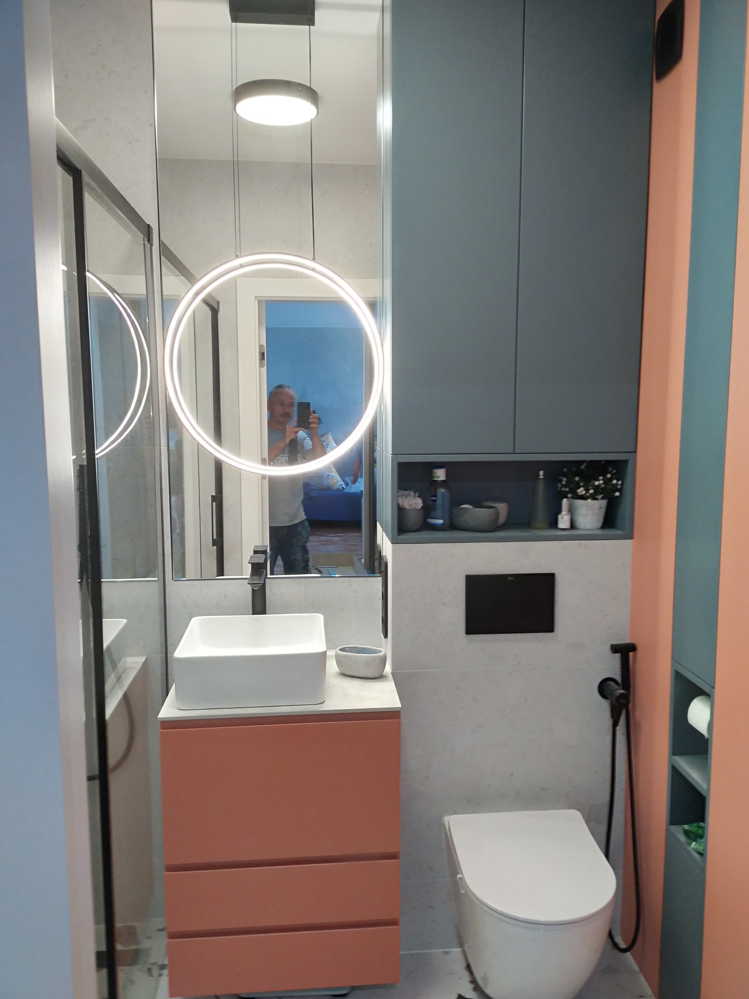 Nowoczesna łazienka z podświetlanym lustrem, umywalką na szafce w odcieniach brzoskwini, wiszącą toaletą i szarą zabudową meblową z otwartą półką z dekoracjami, widok z perspektywy osoby robiącej...