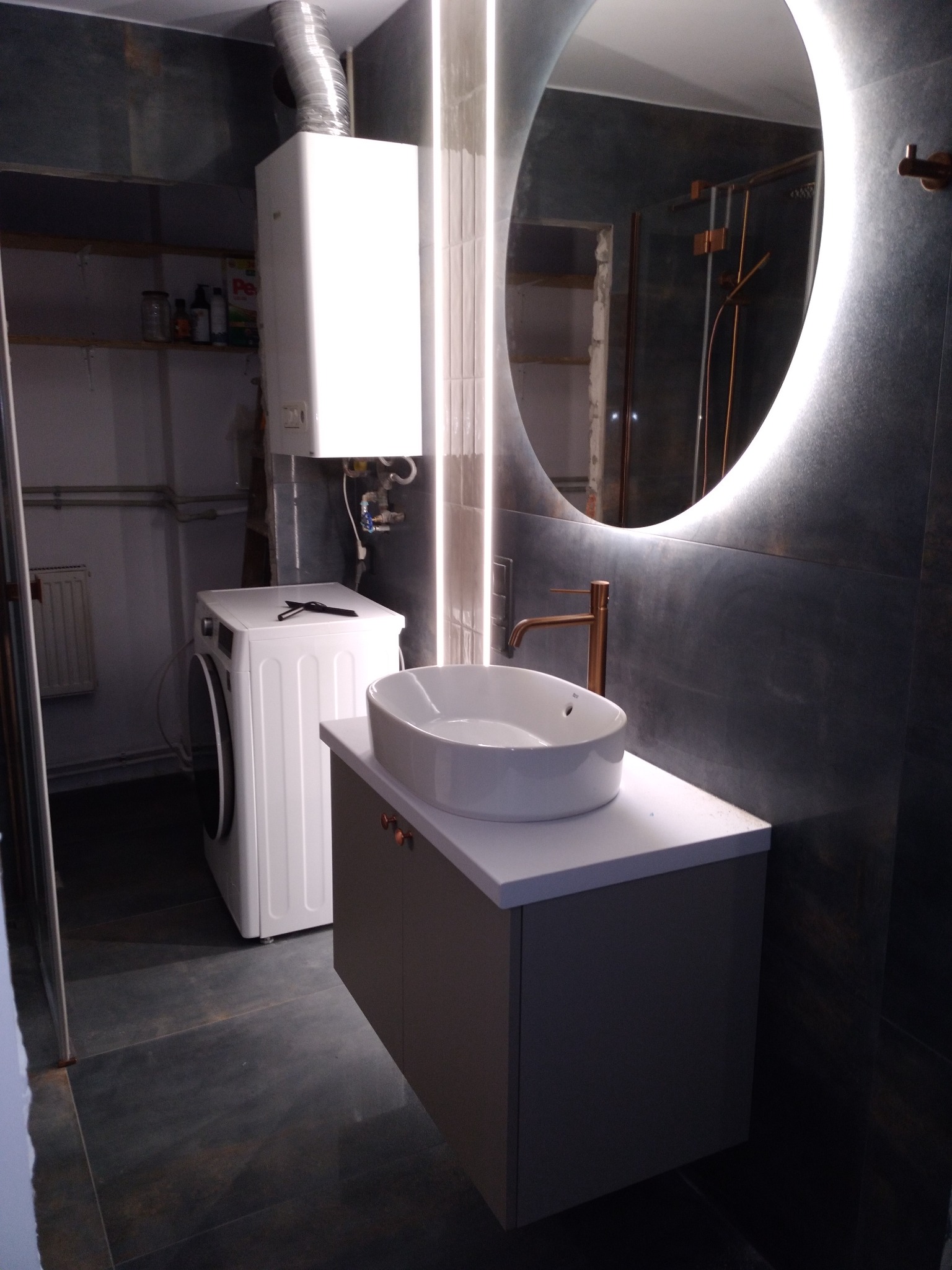 Nowoczesna łazienka z umywalką nablatową, podświetlanym lustrem i pralką w tle; ciemne płytki i minimalistyczny styl.