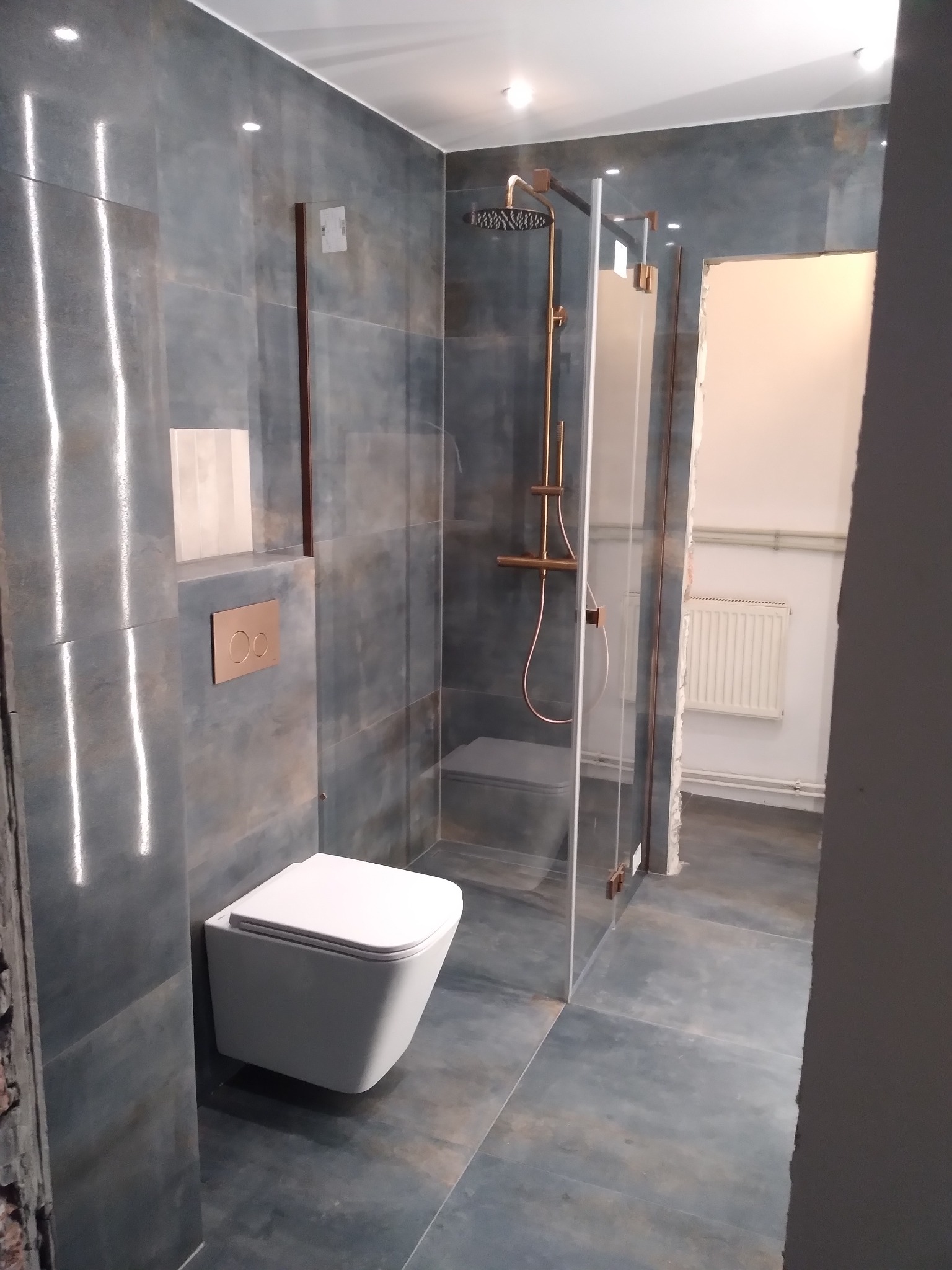 Nowoczesna łazienka z szarymi płytkami imitującymi kamień, podwieszana toaleta, kabina prysznicowa z miedzianą armaturą i widokiem na białe pomieszczenie z grzejnikiem.