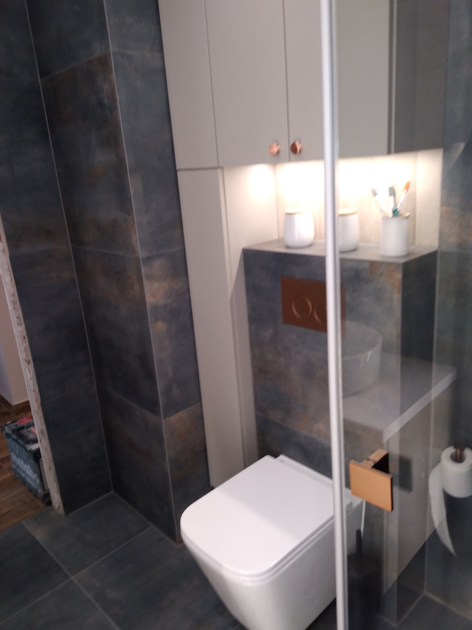 Nowoczesna łazienka z wiszącą toaletą, ciemnymi płytkami imitującymi kamień, zabudową meblową w odcieniach szarości z miedzianymi akcentami i oświetleniem LED nad półką.