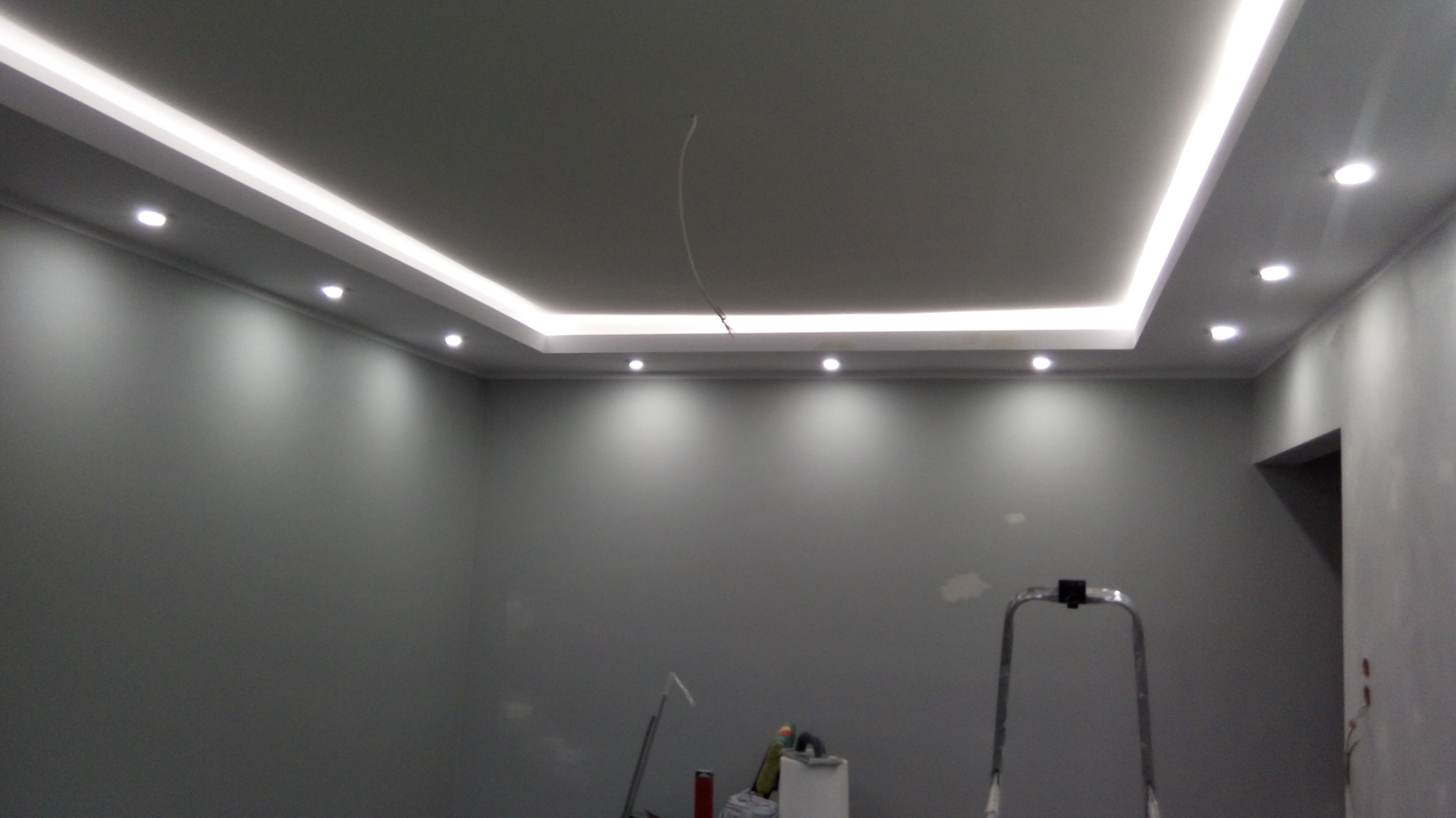 Szare wnętrze po odświeżeniu z podwieszanym sufitem z oświetleniem LED i punktowym, widoczna drabina malarska i kabel zwisający z sufitu.