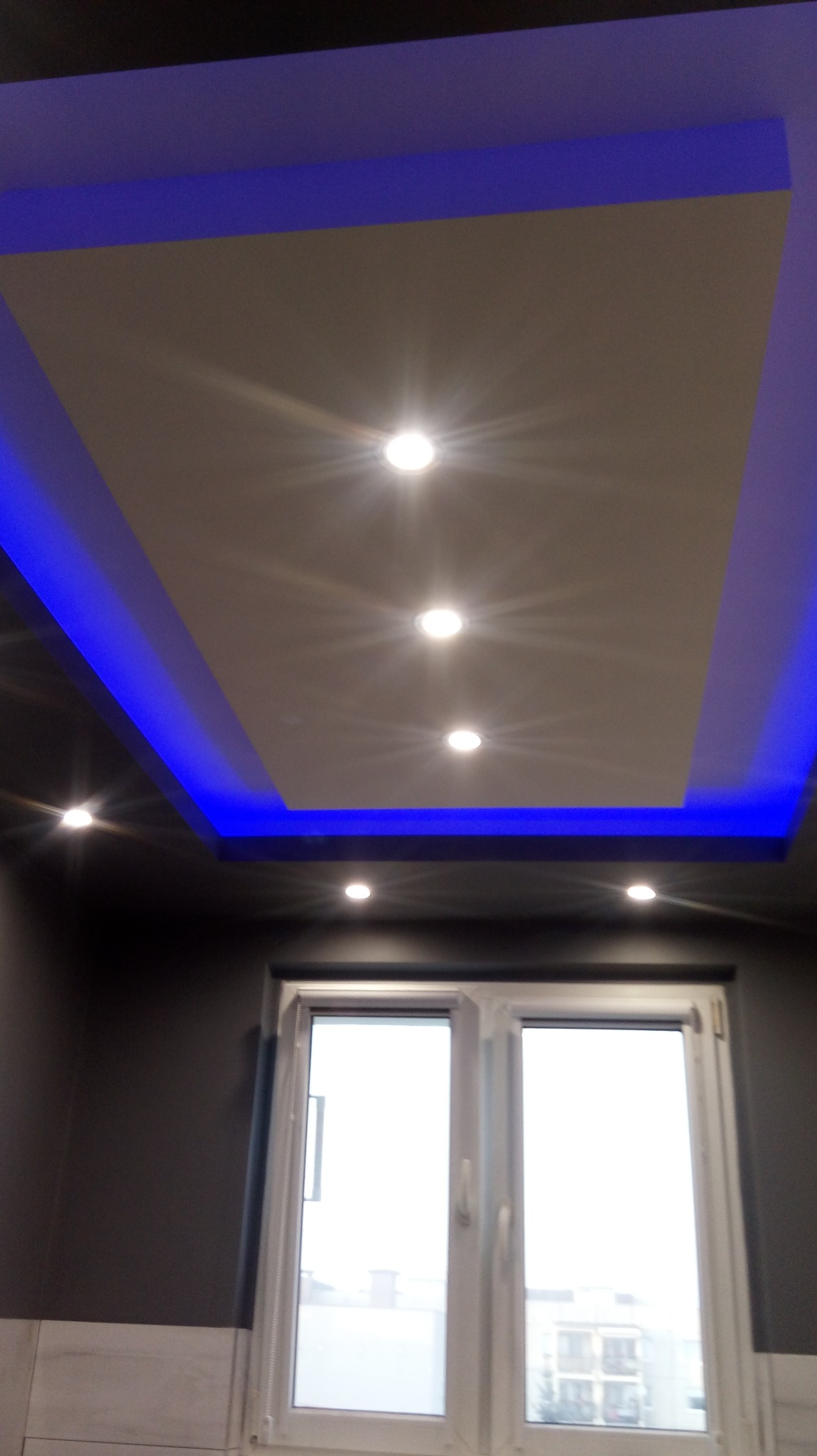 Podwieszany sufit z oświetleniem LED w kolorze niebieskim i centralnie umieszczonymi punktowymi lampami, widok z dołu na tle okna.