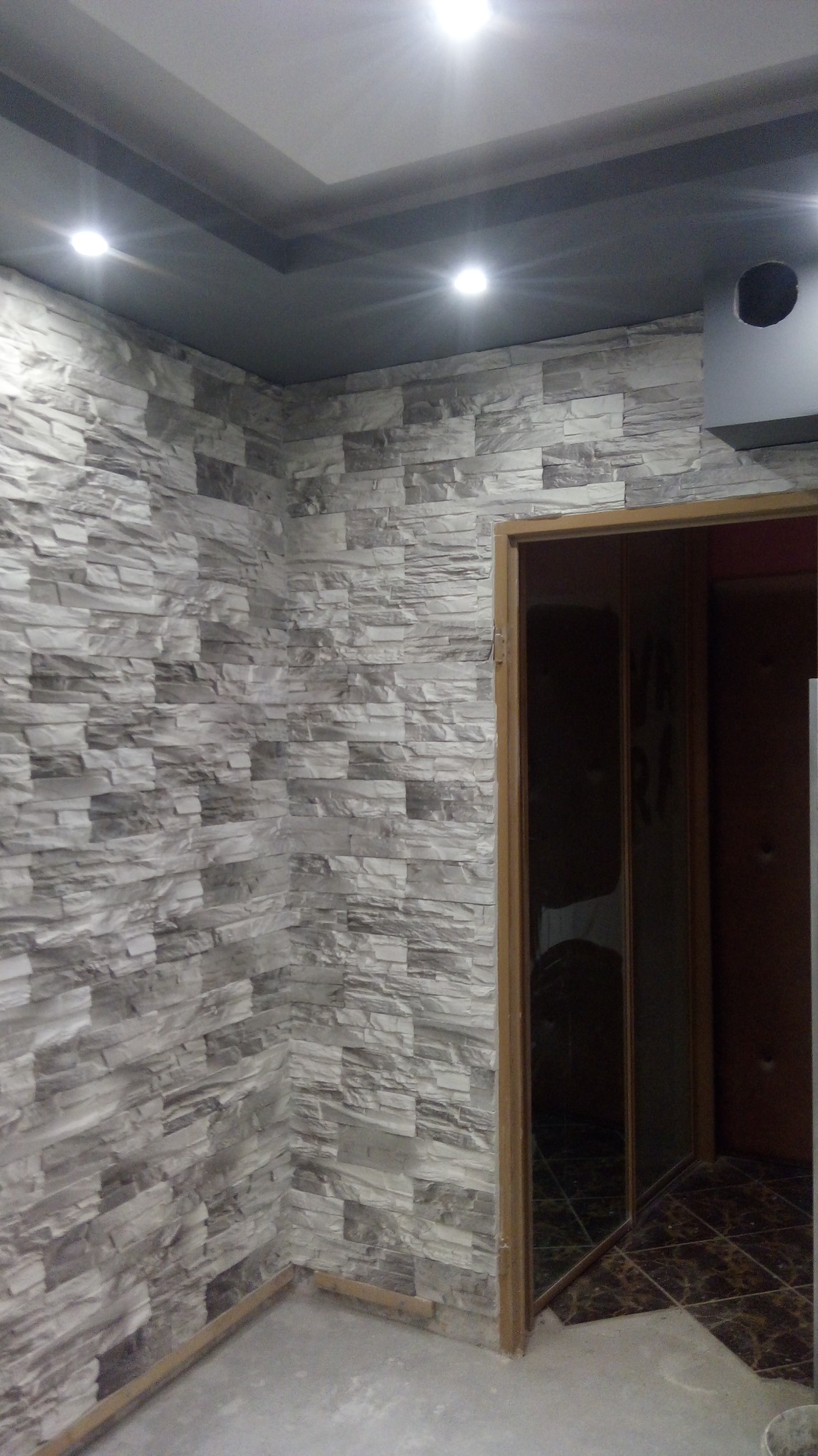 Ściana wykończona panelami imitującymi kamień w odcieniach szarości, oświetlona punktowymi lampami sufitowymi, widok na ościeżnicę drzwiową z fragmentem ciemnej podłogi w tle.