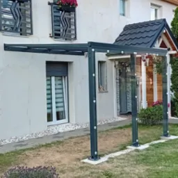 Nowoczesna, metalowa konstrukcja z przezroczystym zadaszeniem i szklanymi ścianami, przylegająca do jasnego domu z balkonami udekorowanymi kwiatami, otoczona trawnikiem i kamiennym podłożem.