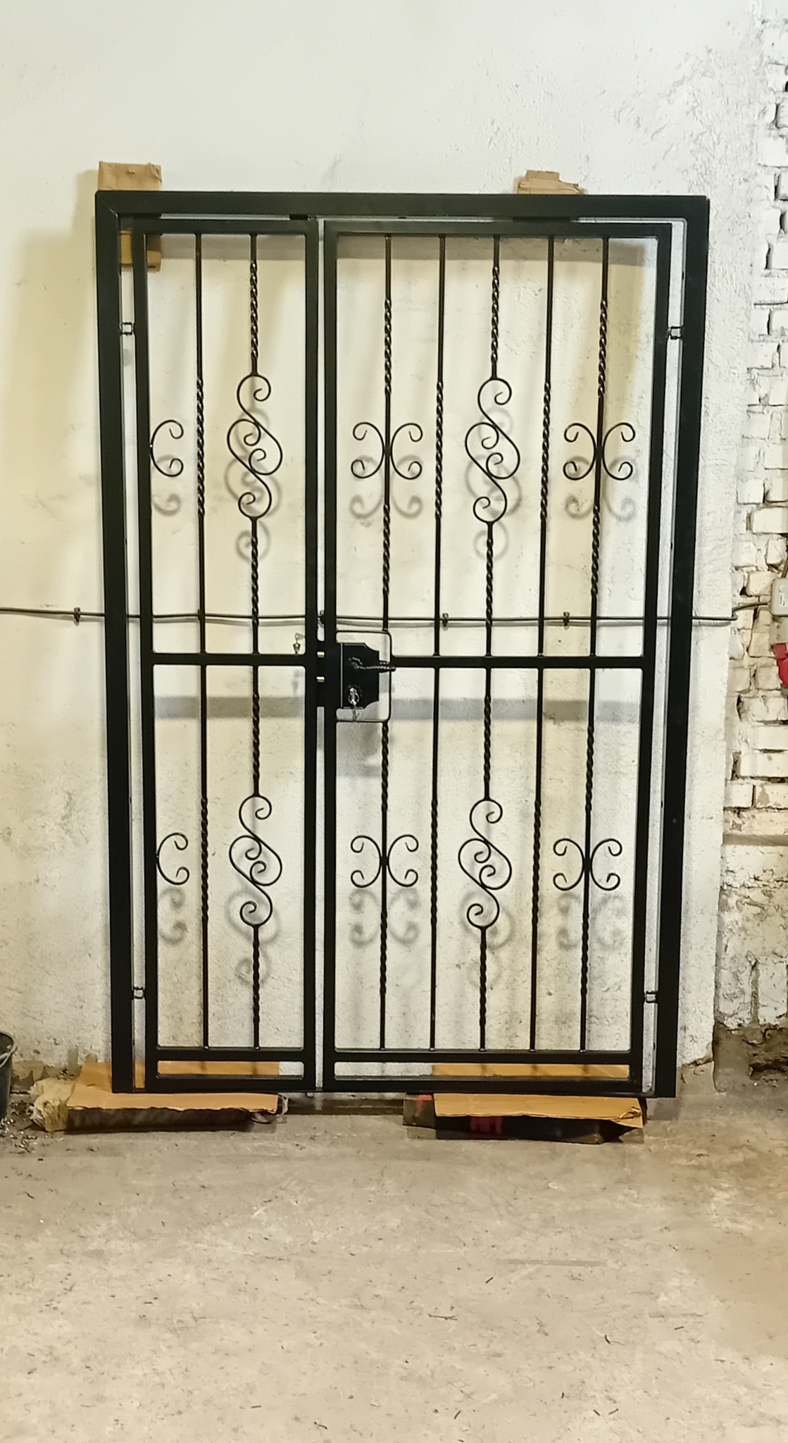 Czarna, metalowa brama dwuskrzydłowa z ozdobnymi elementami, zabezpieczona kartonem, ustawiona na betonowej podłodze przed białą ścianą z fragmentem ściany z białej cegły.