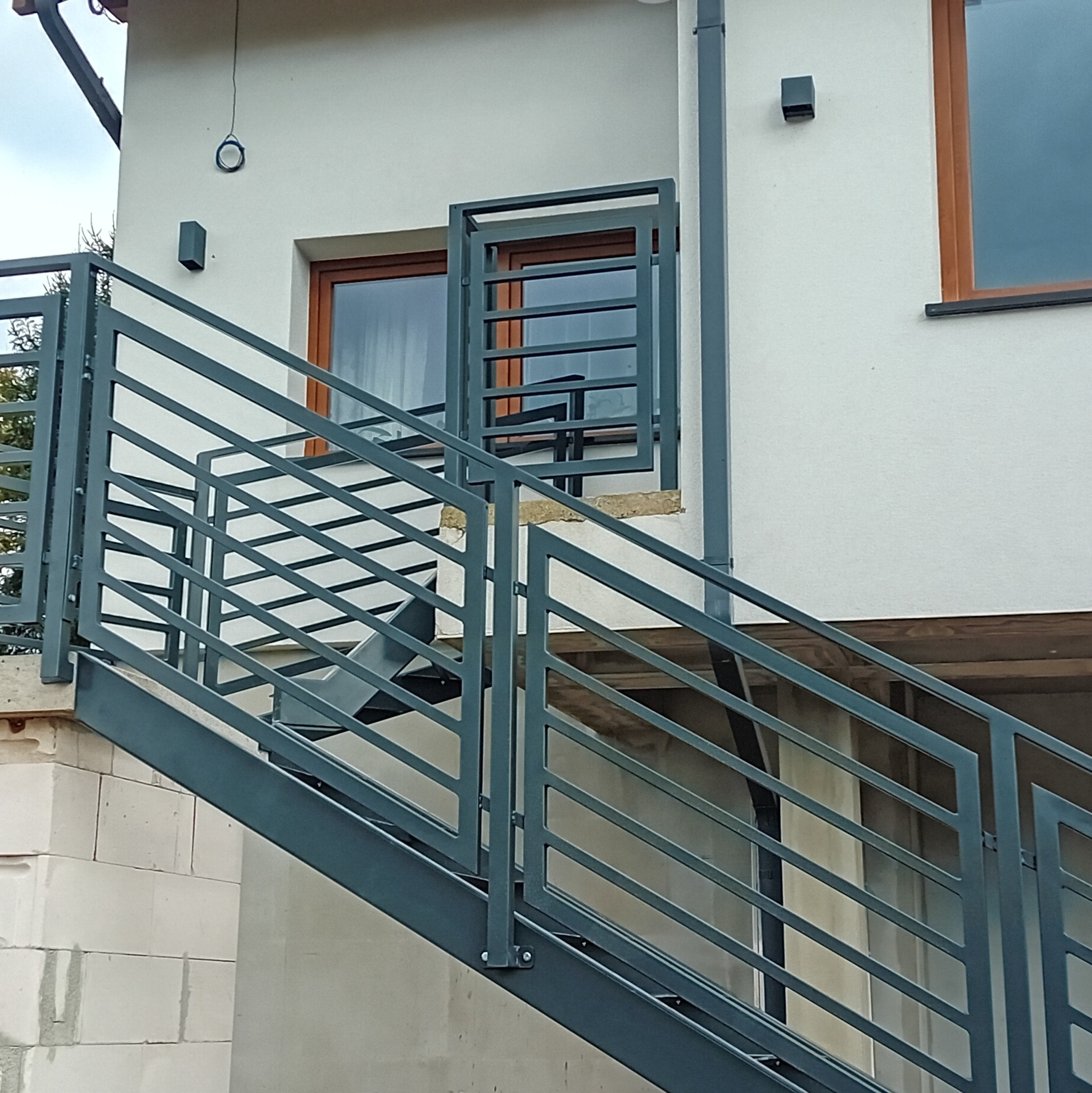 Zewnętrzne schody metalowe z szarą balustradą, prowadzące do drzwi balkonowych na elewacji domu.