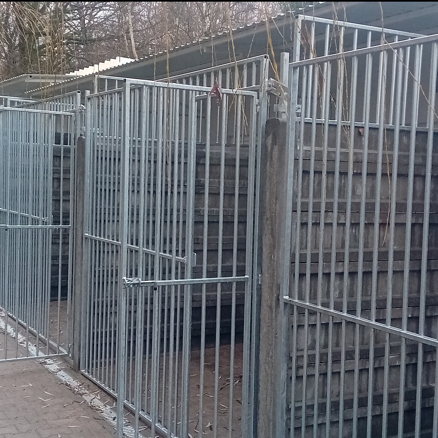 Szereg metalowych, ocynkowanych boksów z kratami i zamykanymi drzwiami w Świerzawie, prawdopodobnie dla zwierząt, z betonowymi słupkami i zadaszeniem w tle.