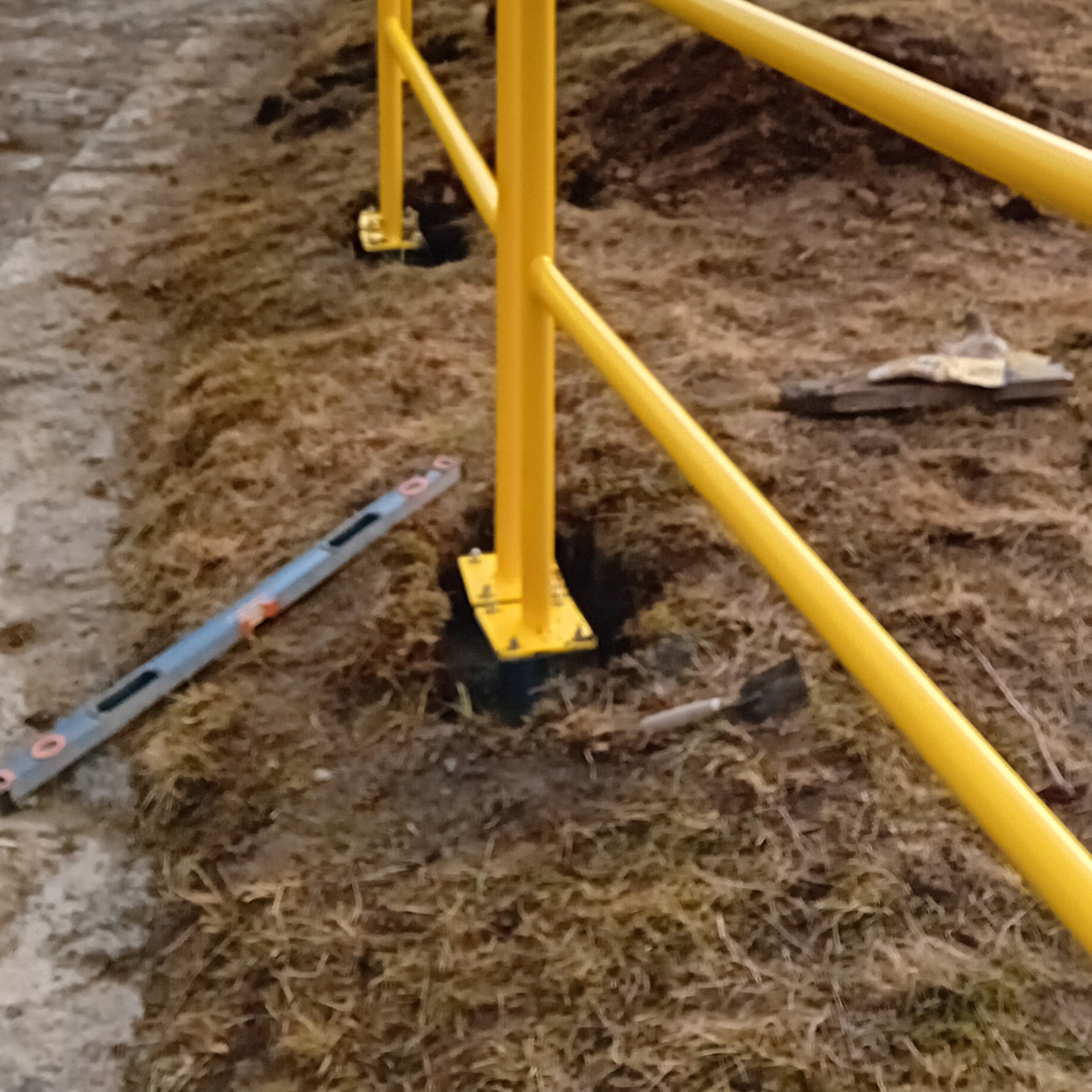 Fragment żółtej metalowej konstrukcji, prawdopodobnie balustrady, z poziomnicą i małą łopatką na ziemi.