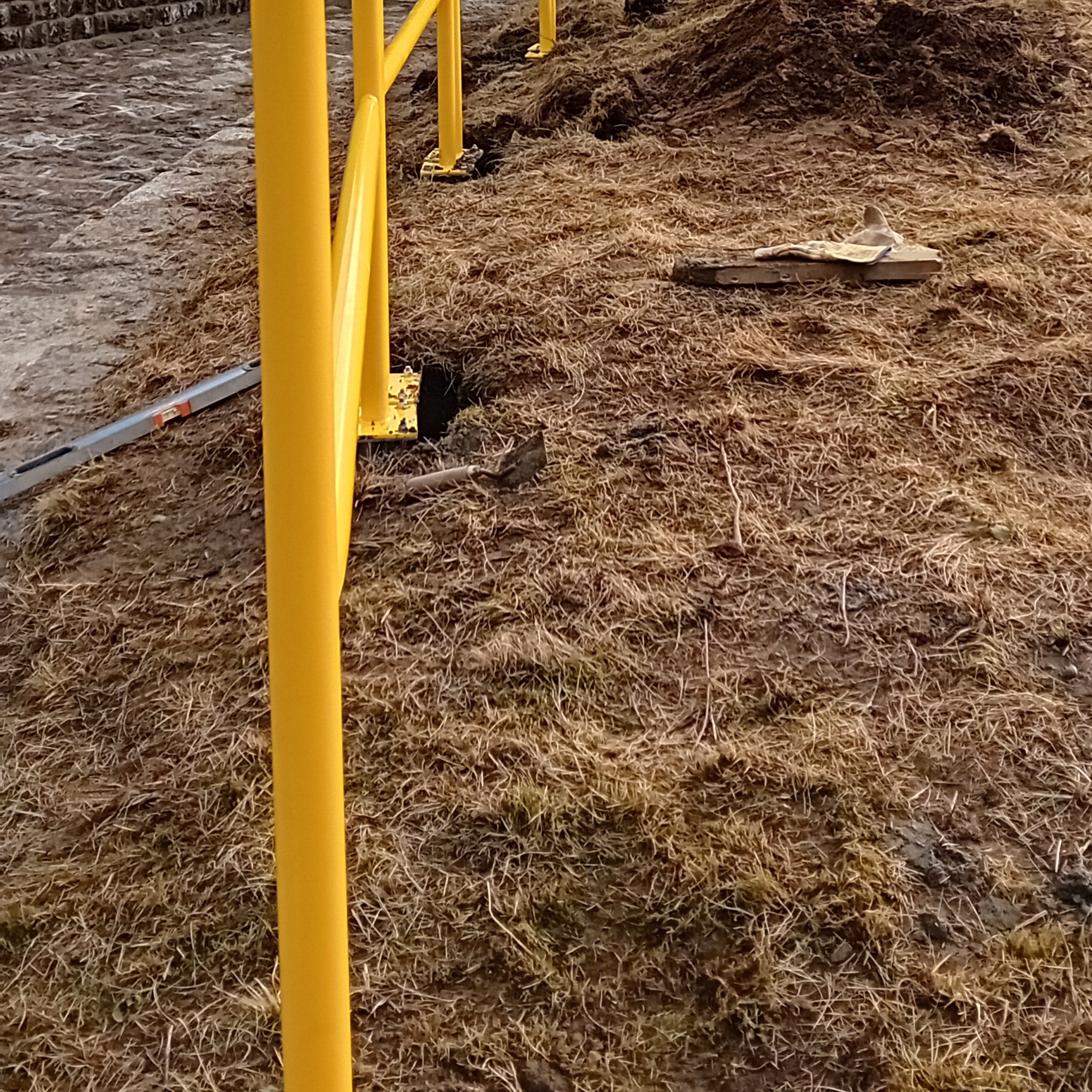 Żółta metalowa konstrukcja w trakcie montażu na zewnątrz, widoczna poziomica, łopata i rękawice robocze na ziemi.