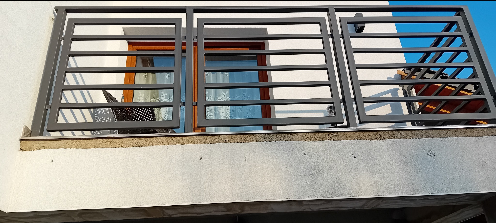 Nowoczesna balustrada balkonowa z szarego metalu o prostokątnych przęsłach, widok z dołu, z fragmentem okna i nieba w tle.