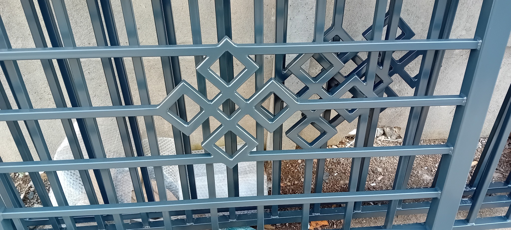 Szczegółowe zbliżenie na fragment metalowej bramy wjazdowej, prezentujący geometryczny wzór złożony z połączonych rombów, pomalowany na szary kolor. Widoczne spawy i konstrukcja kratowa w tle.