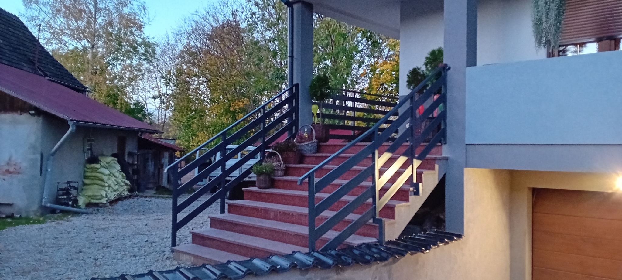 Zewnętrzne schody z czerwonymi stopniami i ciemnymi metalowymi balustradami, prowadzące do zadaszonego wejścia, z dekoracyjnymi koszami i roślinami na stopniach.