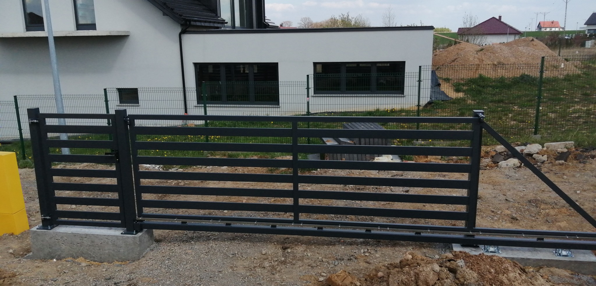 Szara, metalowa brama przesuwna z poziomymi szczeblami, zamontowana na betonowej podstawie przed nowym domem w trakcie budowy.