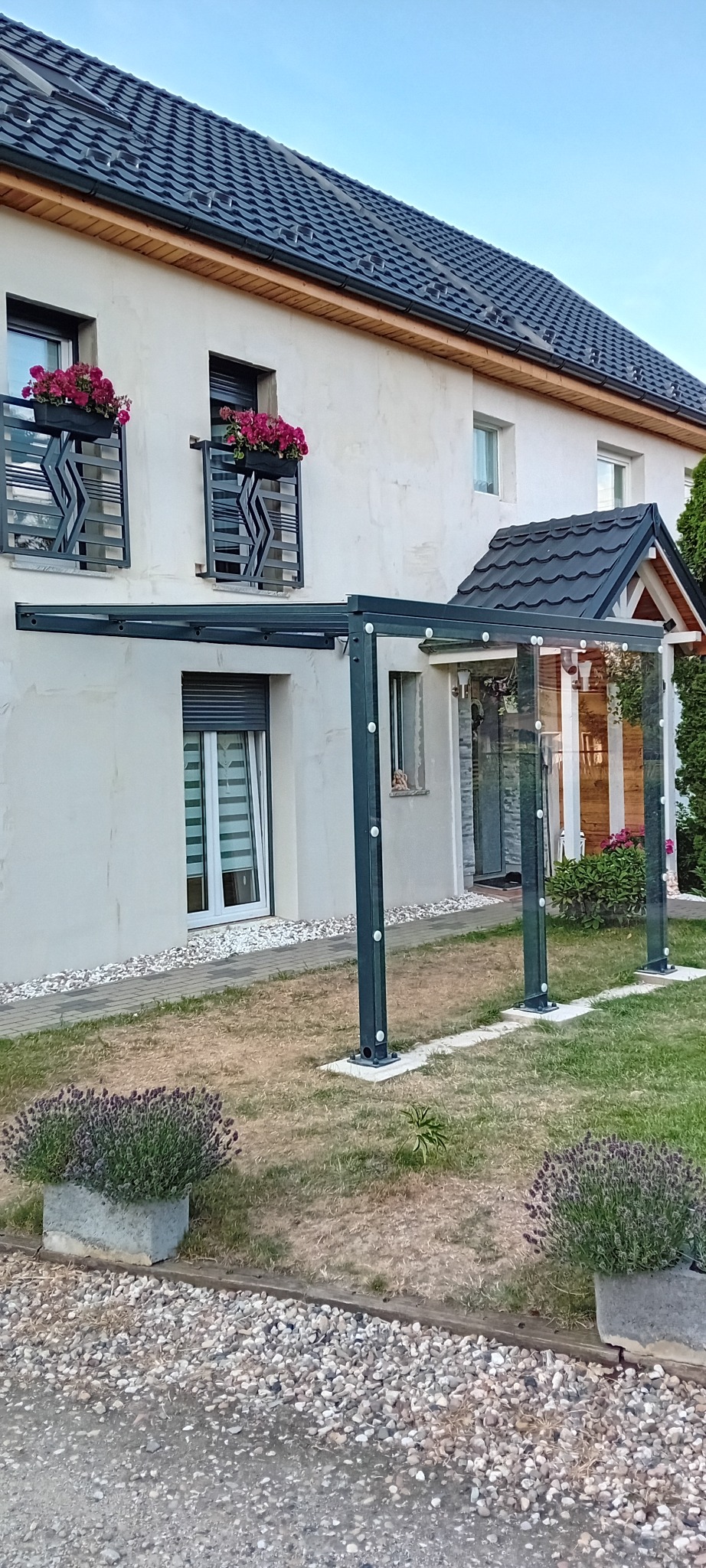 Nowoczesna, metalowa konstrukcja z przezroczystym zadaszeniem i szklanymi ścianami, przylegająca do jasnego domu z balkonami udekorowanymi kwiatami, otoczona trawnikiem i kamiennym podłożem.