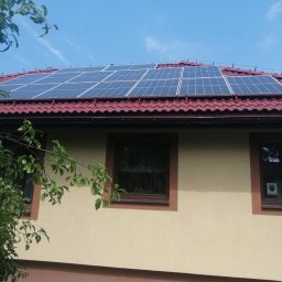 EkoOszczędni - Panele słoneczne na dachu domu z czerwoną dachówką, widoczne z poziomu gruntu, z fragmentem drzewa po lewej stronie.