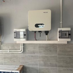 EkoOszczędni - Zamontowany inwerter Sofar z przewodami i skrzynkami zabezpieczeń na tle ściany wyłożonej płytkami imitującymi drewno, z widocznymi panelami fotowoltaicznymi w dolnej części kadru.