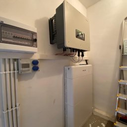 EkoOszczędni - Biała bateria magazynu energii Sofar i szary inwerter fotowoltaiczny zamontowane na ścianie w pomieszczeniu gospodarczym, obok skrzynki elektrycznej i drabiny.