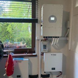 EkoOszczędni - Wnętrze z zamontowanym inwerterem i magazynem energii V-TAC na ścianie, widok z okna z roletami i zielenią na zewnątrz.