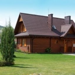 EkoOszczędni - Gotowy, drewniany dom z brązowym dachem, kominem i jasną elewacją na tle zielonego trawnika i gór.