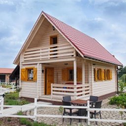 EkoOszczędni - Gotowy, dwukondygnacyjny dom szkieletowy z czerwonym dachem, drewnianymi okiennicami i balkonem, otoczony białym płotem i zadbanym ogrodem.