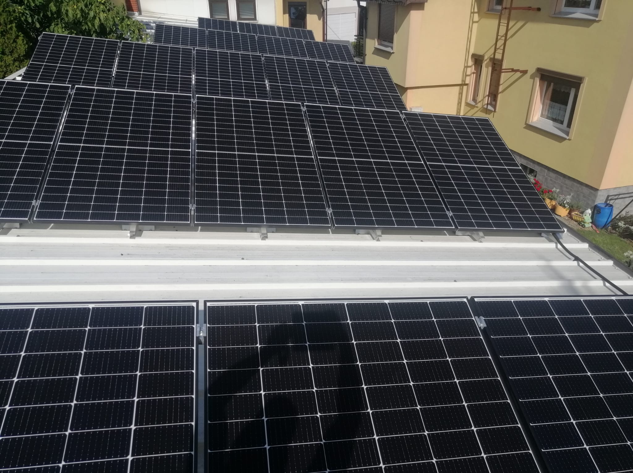 Panele fotowoltaiczne zamontowane na białym dachu budynku mieszkalnego, widoczne okna i drabina na elewacji.