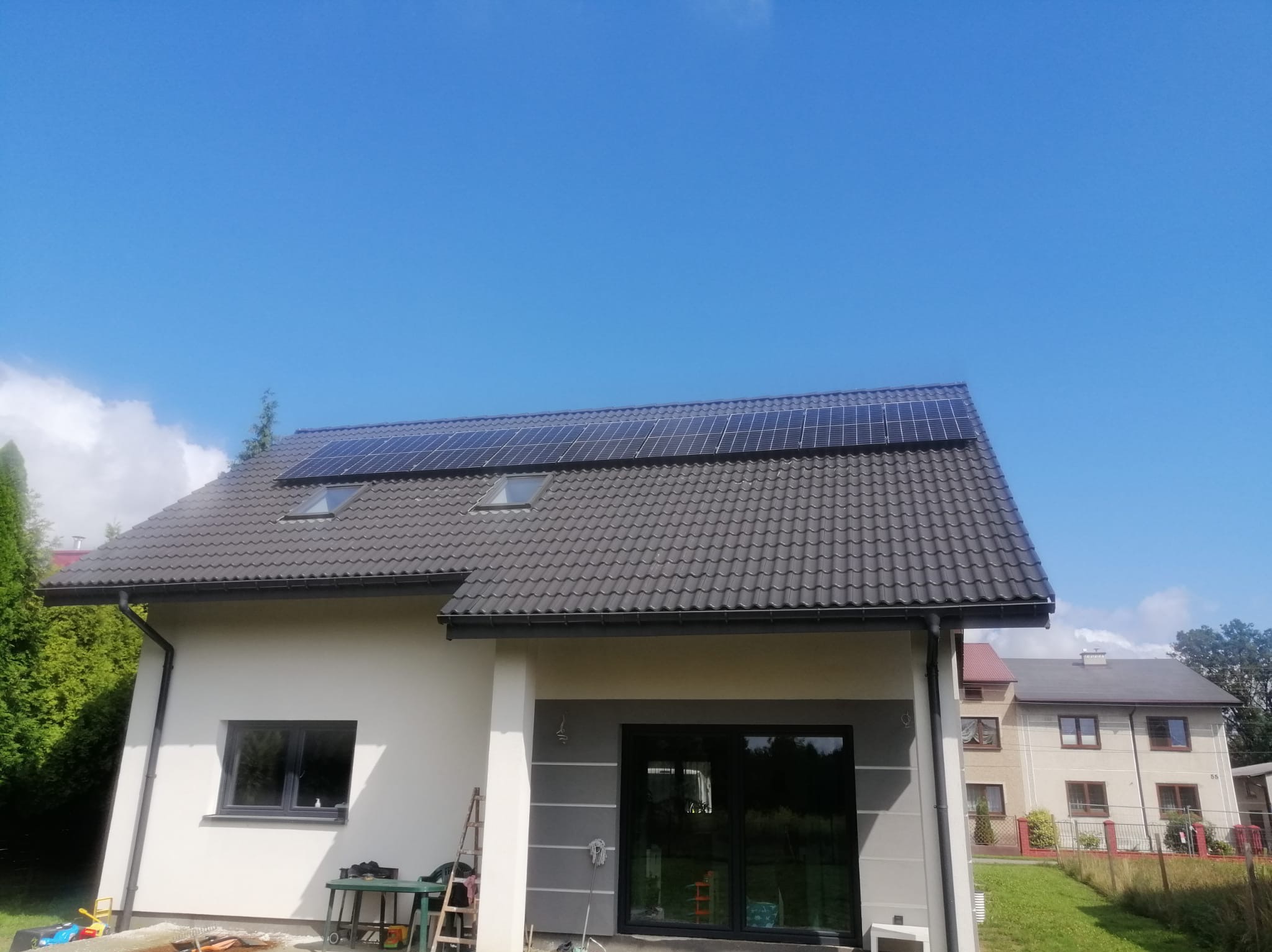 Nowoczesny dom jednorodzinny z szarym dachem i panelami słonecznymi, widok z dołu na tle błękitnego nieba, z widocznymi oknami dachowymi i elementami elewacji w odcieniach bieli i szarości.