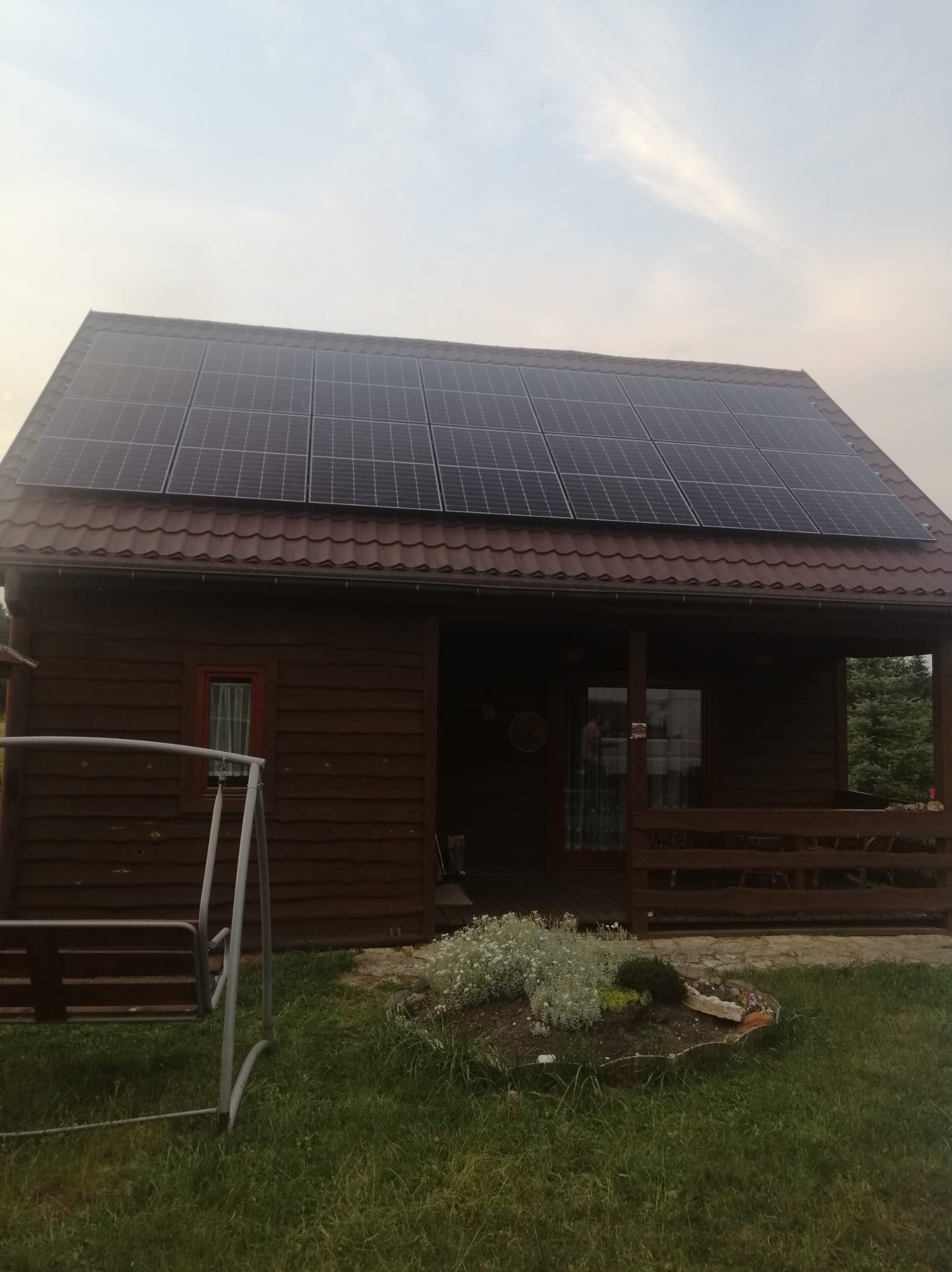 Drewniany domek z brązowym dachem pokrytym panelami słonecznymi. Na pierwszym planie huśtawka i rabata z kwiatami.