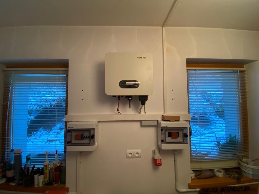 Zamontowany inwerter SOFAR na białej ścianie pomiędzy dwoma oknami z żaluzjami, poniżej widoczne dwie skrzynki elektryczne i gniazdko, okablowanie.