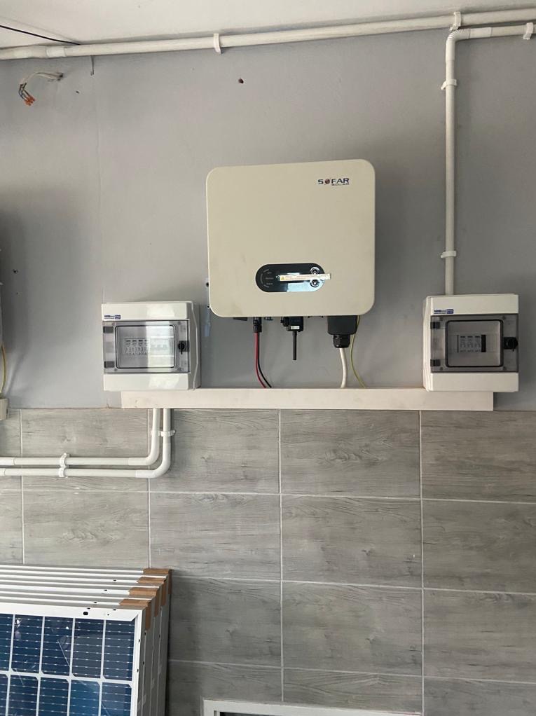 Zamontowany inwerter Sofar z przewodami i skrzynkami zabezpieczeń na tle ściany wyłożonej płytkami imitującymi drewno, z widocznymi panelami fotowoltaicznymi w dolnej części kadru.