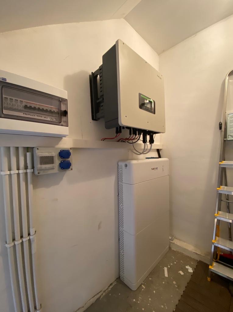 Biała bateria magazynu energii Sofar i szary inwerter fotowoltaiczny zamontowane na ścianie w pomieszczeniu gospodarczym, obok skrzynki elektrycznej i drabiny.