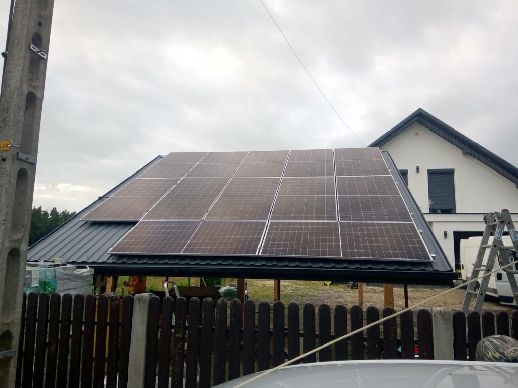 Panele słoneczne zamontowane na dachu wiaty, widok zza drewnianego płotu, w tle dom jednorodzinny z drabina oparta o sciane.