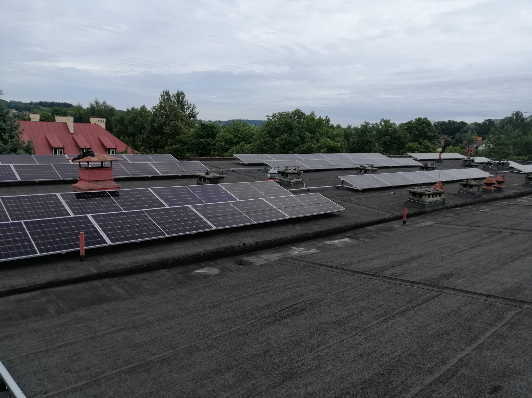 Panele słoneczne na płaskim dachu budynku z kominami, widok z góry na tle zieleni i pochmurnego nieba.