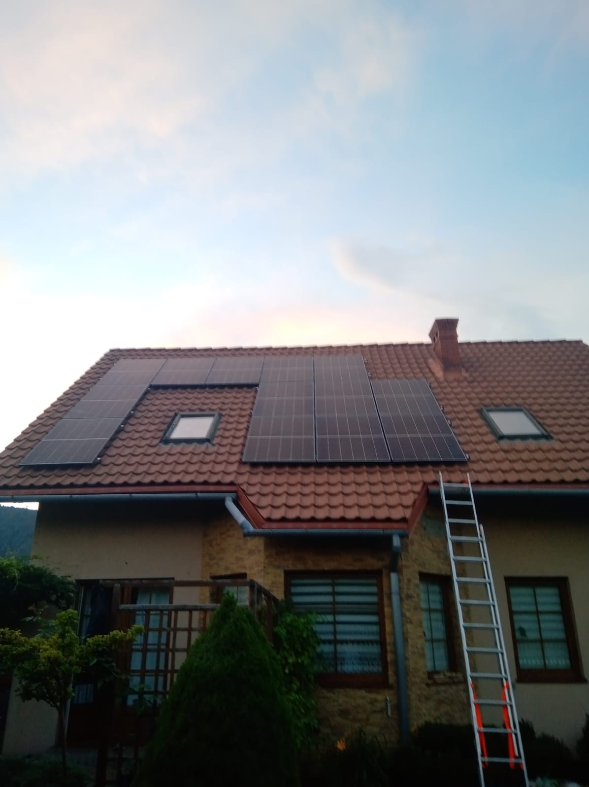 Panele słoneczne zamontowane na dachu domu z czerwoną dachówką, z widoczną drabiną opartą o ścianę budynku.