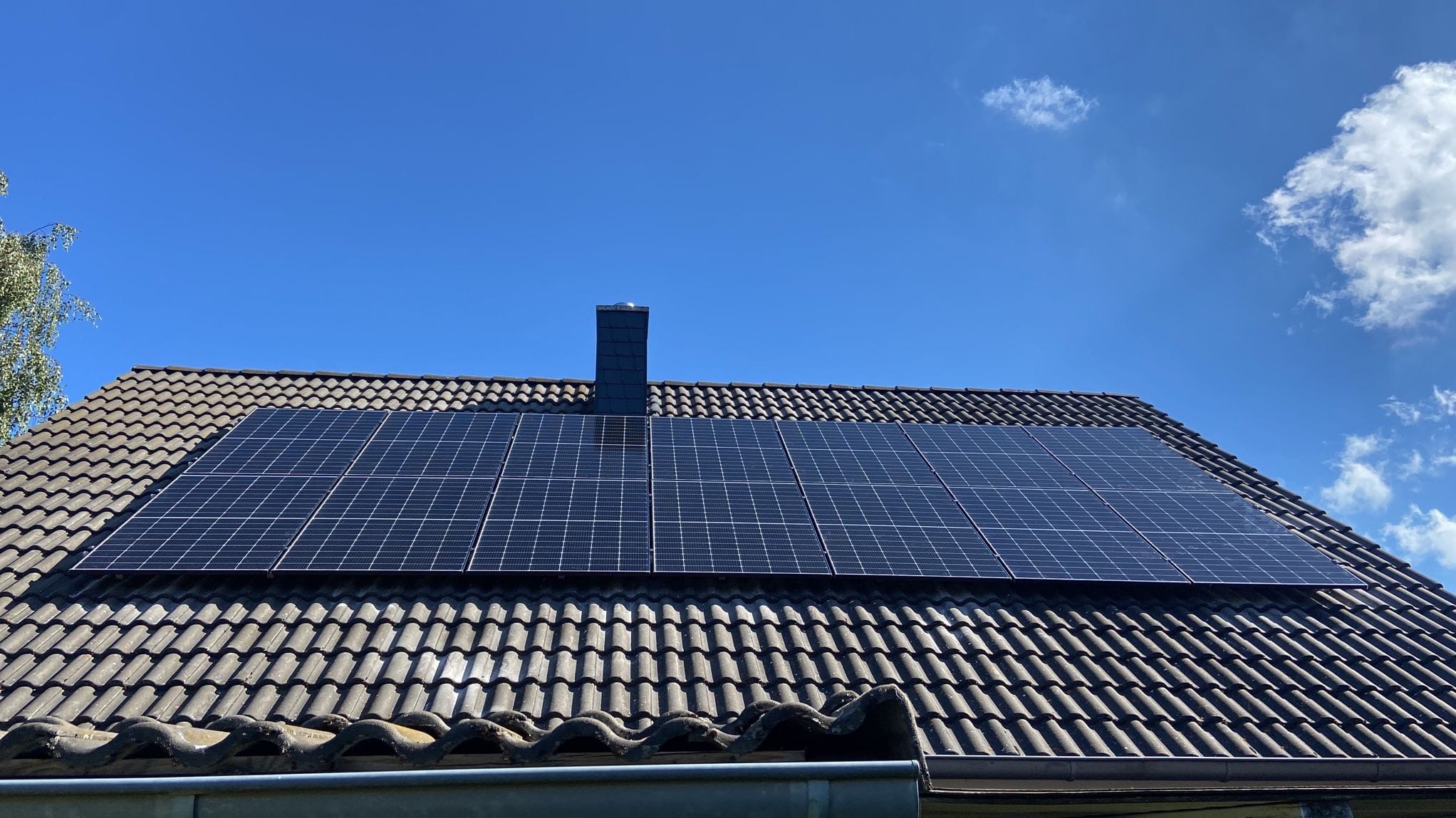 Panel słoneczny na dachu z dachówką ceramiczną, komin w tle, błękitne niebo z chmurami.