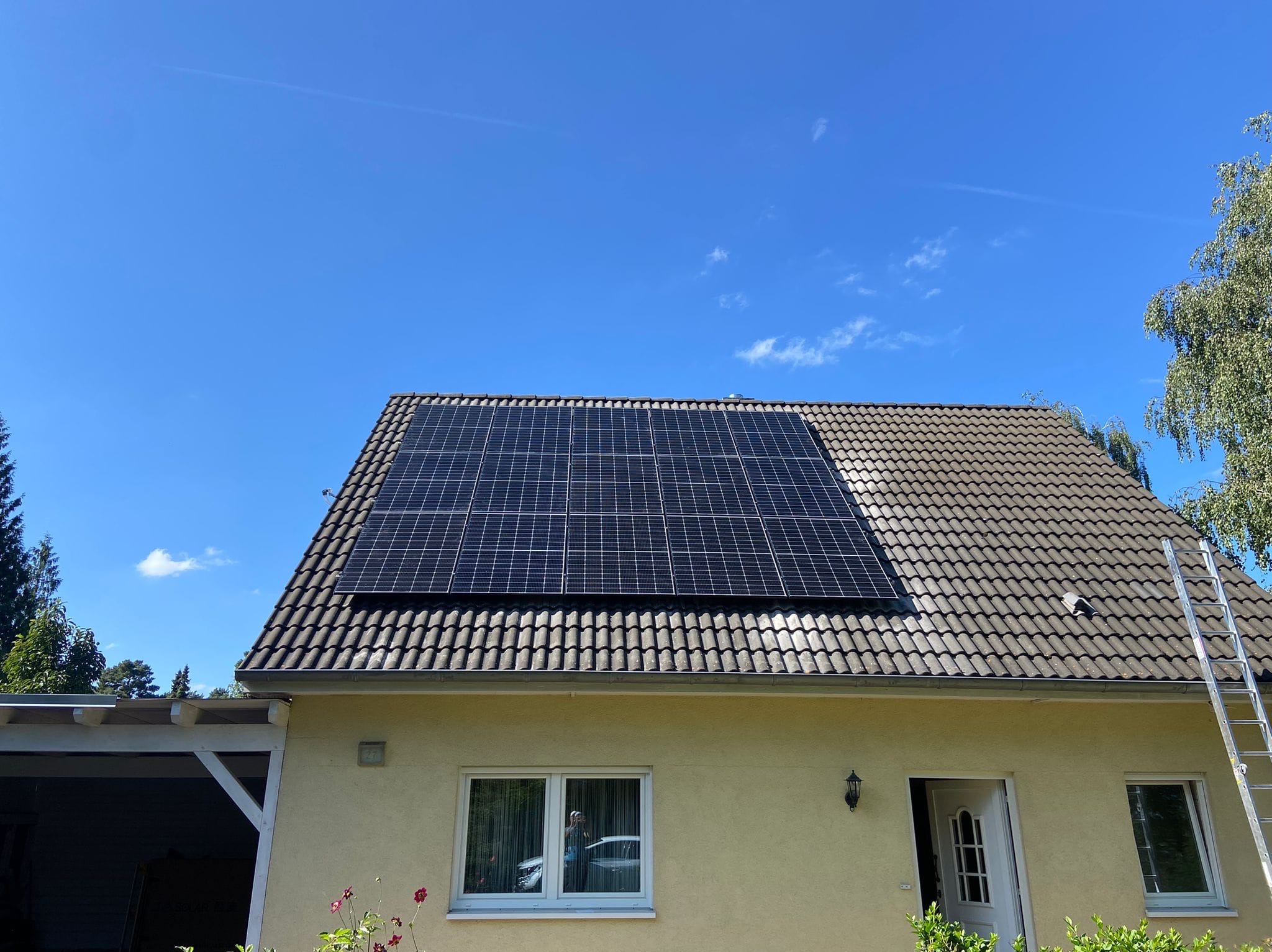 Instalacja fotowoltaiczna na dachu domu jednorodzinnego z drabina opartą o ścianę budynku, widok z dołu w słoneczny dzień.