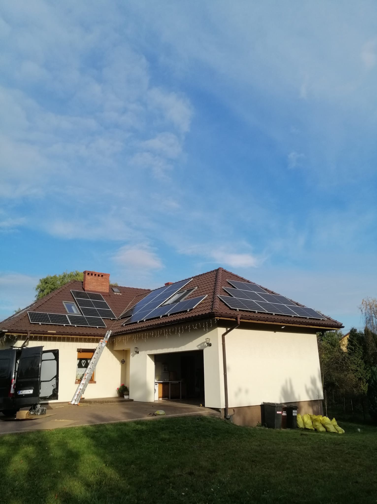 Instalacja paneli fotowoltaicznych na dachu domu z brązową dachówką, widoczna drabina oparta o ścianę, samochód dostawczy z otwartymi drzwiami i worki na trawniku, błękitne niebo w tle.