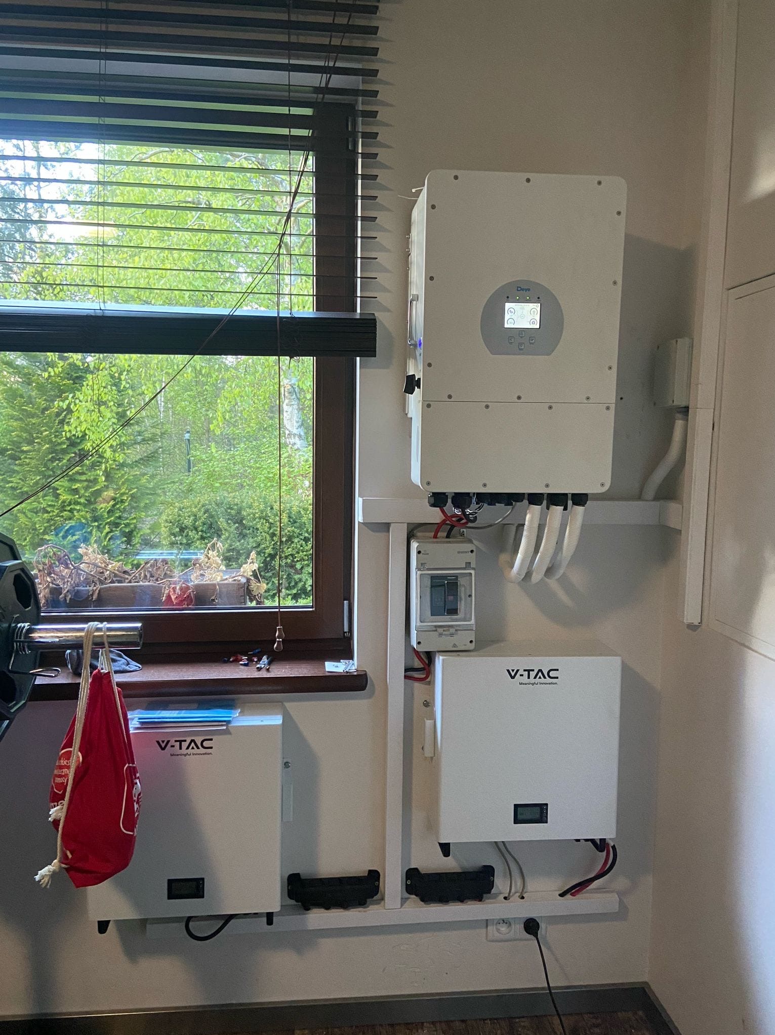 Wnętrze z zamontowanym inwerterem i magazynem energii V-TAC na ścianie, widok z okna z roletami i zielenią na zewnątrz.