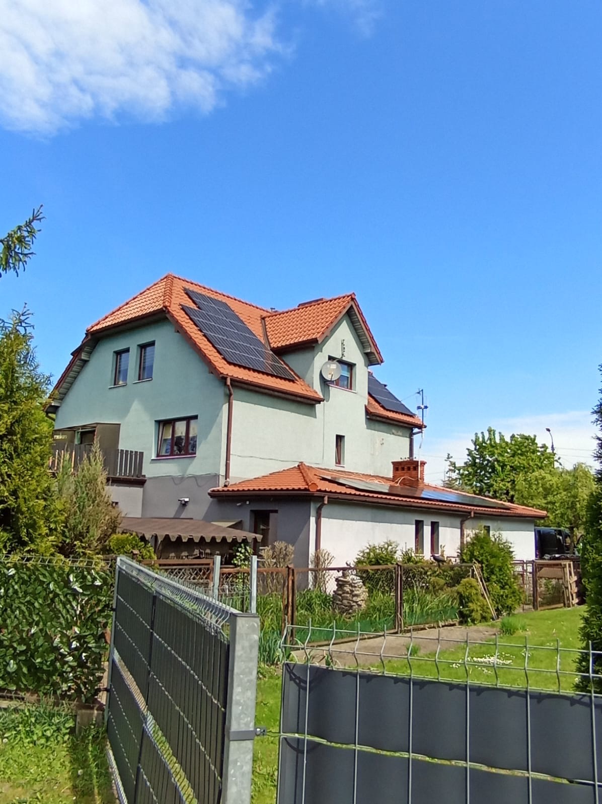 Dwukondygnacyjny dom z pomalowaną na szaro elewacją i pomarańczową dachówką, na której zamontowano panele słoneczne; widok zza ogrodzenia w słoneczny dzień.