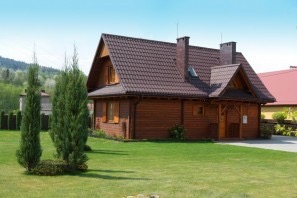 Gotowy, drewniany dom z brązowym dachem, kominem i jasną elewacją na tle zielonego trawnika i gór.