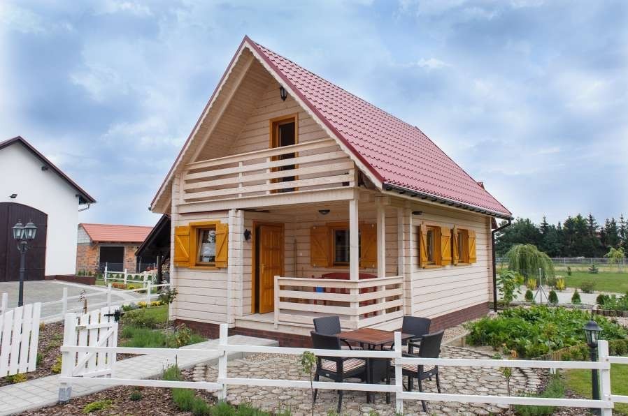 Gotowy, dwukondygnacyjny dom szkieletowy z czerwonym dachem, drewnianymi okiennicami i balkonem, otoczony białym płotem i zadbanym ogrodem.