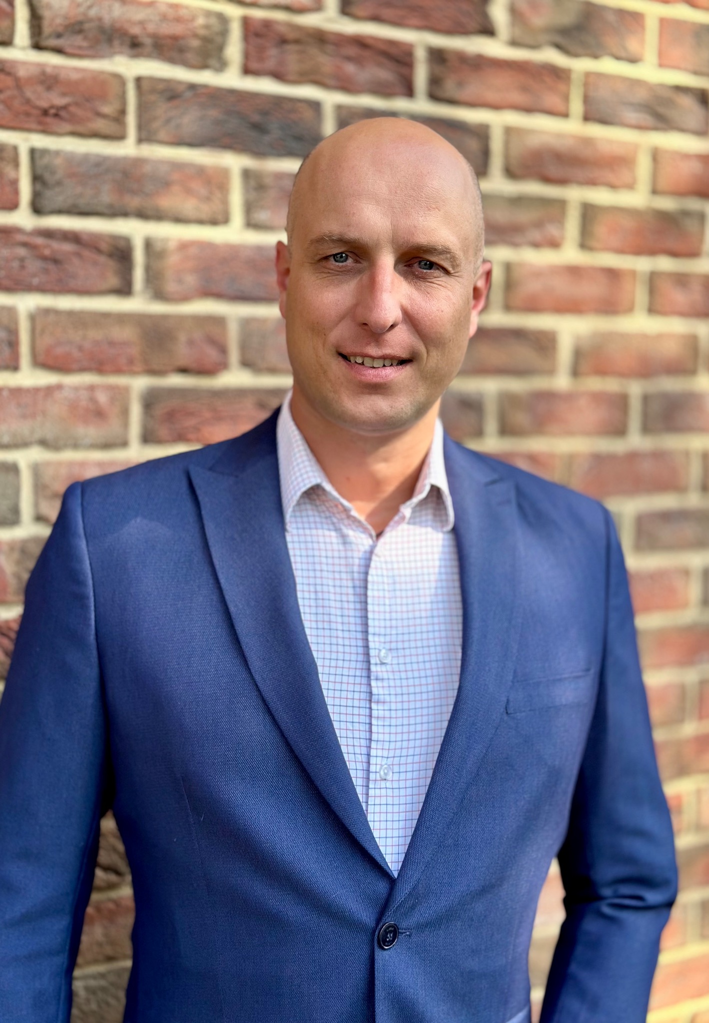 Portret uśmiechniętego mężczyzny w niebieskiej marynarce i koszuli w kratkę, stojącego na tle ściany z czerwonej cegły.