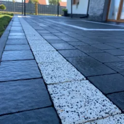 Nowo ułożona kostka brukowa w kolorze grafitowym z białym pasem dekoracyjnym z drobnych kamyczków, widok z niskiej perspektywy na tle domu.